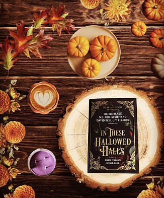 photograph of a copy of In These Hallowed Halls, edited by Marie O'Regan and Paul Kane, lying on a log slice on a wooden table. Orange flowers and leaves edge the image, and beside the log are a purple three-wicked jar candle, a coffee with a heart design on top, and three small pumpkins on a beige plate