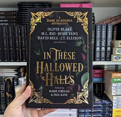Image of a hand holding up a copy of In These Hallowed Halls, edited by Marie O'Regan and Paul Kane, against a background of filled bookshelves at the Titan offices