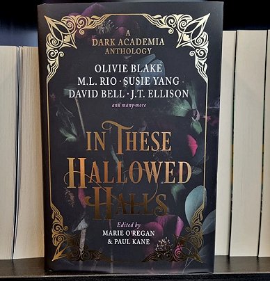 photograph of a copy of In These Hallowed Halls, edited by Marie O'Regan and Paul Kane, standing on a shelf in front of a row of books, page edges facing out