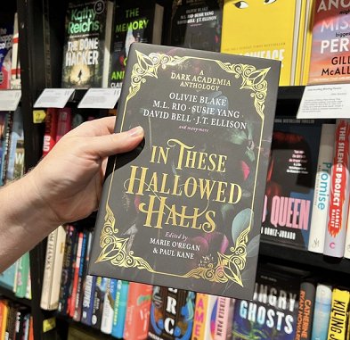 photograph of a hand holding a copy of In These Hallowed Halls, edited by Marie O'Regan and Paul Kane, up against shelves in a bookstore