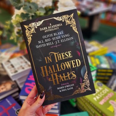 woman's hand holds a copy of In These Hallowed Halls, edited by Marie O'Regan and Paul Kane, over a book piled high with books