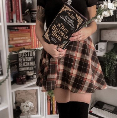 photograph of a woman in a brown plaid skirt, black stockings and black top, standing in front of packed bookshelves and holding a copy of In These Hallowed Halls, edited by Marie O'Regan and Paul Kane