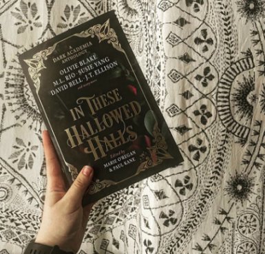 a hand holds a copy of In These Hallowed Halls, edited by Marie O'Regan and Paul Kane, above a white bedspread with a complicated black embroidered pattern