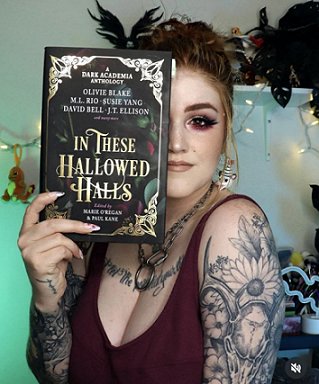 photograph of a young woman with tattoos on her arms and chest, holding up a copy of In These Hallowed Halls, edited by Marie O'Regan and Paul Kane