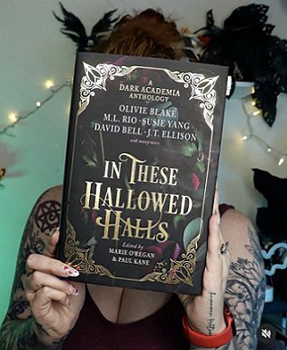 photograph of a young woman with tattoos on her arms, holding a copy of In These Hallowed Halls, edited by Marie O'Regan and Paul Kane, up in front of her face