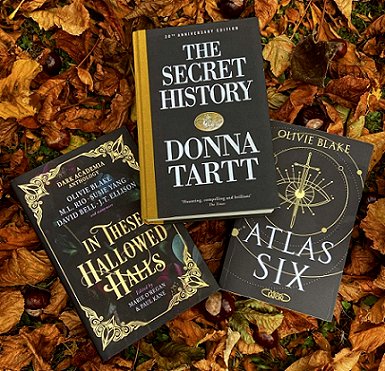 Three books lying on gold and brown autumn leaves. The books are: In These Hallowed Halls, edited by Marie O'Regan and Paul Kane, The Secret History by Donna Tartt, and Atlas Six by Olivie Blake