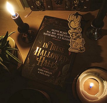 photograph showing a copy of In These Hallowed Halls, edited by Marie O'Regan and Paul Kane, lying on a wooden surface - the image is candlelit, and the book is bordered with a plant, two candles and a bookmark