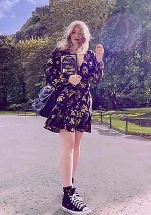 photograph of a woman with windswept long blonde hair wearing black Converse and a dark floral dress and handbag, holding a copy of In These Hallowed Halls, edited by Marie O'Regan and Paul Kane. In the background are grass, trees, and a rocky hillside