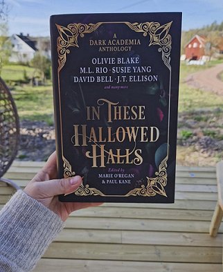 photograph of a woman's hand holding a copy of In These Hallowed Halls, edited by Marie O'Regan and Paul Kane, up against a background of fields, with a white and a red house further back, as well as trees and a blue sky