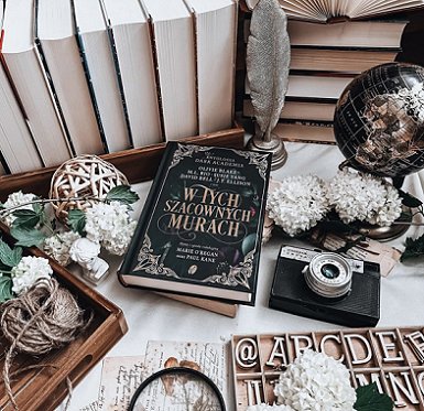 photograph of a copy of the Polish edition of In These Hallowed Halls, edited by Marie O'Regan and Paul Kane, lying on a white surface, surrounded by white flowers, a camera, white letter stencils, twine balls, a globe, a silver quill and holder, and several hardback books standing page side out