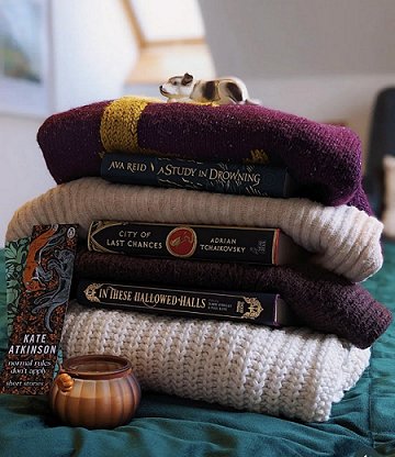 photograph of a pile of folded jumpers in cream and burgundy on a green quilt. In this pile are copies of A Study in Drowning by Ava Reid, City of Last Chances by Adrian Tchaikovsky and In These Hallowed Halls, edited by Marie O'Regan and Paul Kane. In front is a pumpkin-shaped glass jar candle and a Kate Atkinson bookmark wtih a fox on the front