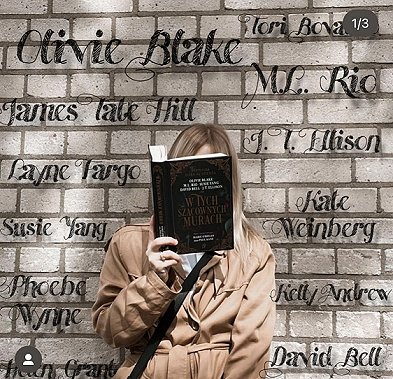 photograph of a blonde woman in a beige coat holding up a copy of the Polish edition of In These Hallowed Halls, edited by Marie O'Regan and Paul Kane. The woman is standing in front of a brick wall featuring the names of all the contributors