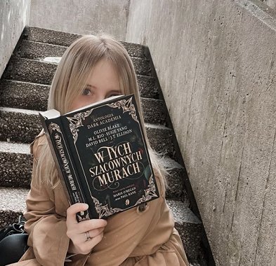 photograph of a young blonde woman in a beige coat sitting on concrete steps, holding up a copy of the Polish edition of In These Hallowed Halls, edited by Marie O'Regan and Paul Kane