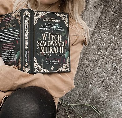 photograph of a young blonde woman in a beige coat sitting against a concrete wall, holding an open copy of the Polish edition of In These Hallowed Halls, edited by Marie O'Regan and Paul Kane