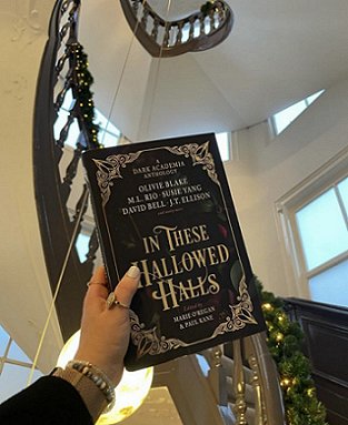 photograph of a woman's hand holding a copy of In These Hallowed Halls, edited by Marie O'Regan and Paul Kane, up against a black spiral staircase