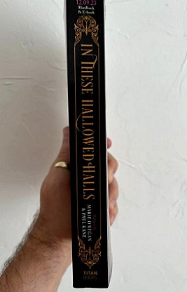A man's hand holding a copy of In These Hallowed Halls, edited by Marie O'Regan and Paul Kane, so that the spine of the book is showing
