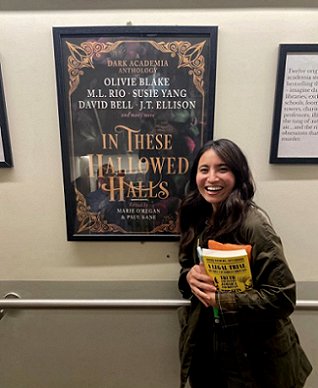 Olivie Blake standing in front of a poster showing the cover of In These Hallowed Halls, edited by Marie O'Regan and Paul Kane