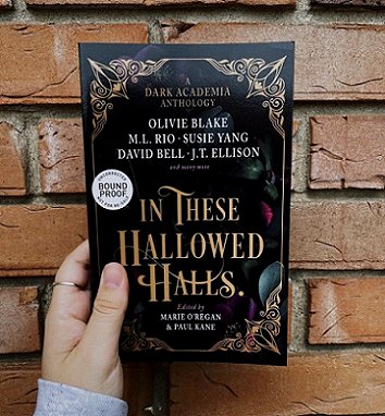 Image of a woman's hand holding a copy of In These Hallowed Halls, edited by Marie O'Regan and Paul Kane, up against a brick wall