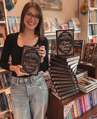 photograph of author Kelly Andrew wearing a black long-sleeved top and blue jeans, holding a copy of In These Hallowed Halls, edited by Marie O'Regan and Paul Kane. Nex to Kelly is a stack of copies of the book, with one standing tall on top