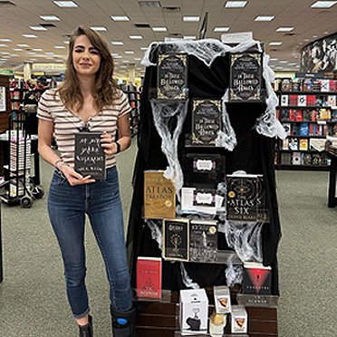Author M L Rio standing beside a display featuring dark academia books including In These Hallowed Halls, edited by Marie O'Regan and Paul Kane. She's holding a copy of her novel, If We Were Villains