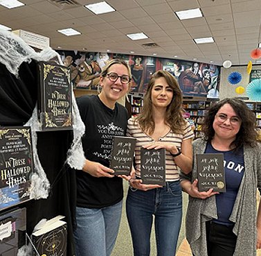 photograph of M L Rio standing between two women, one blonde, one dark, all holding copies of If We Were Villains. Beside them is a display featuring In These Hallowed Halls, edited by Marie O'Regan and Paul Kane