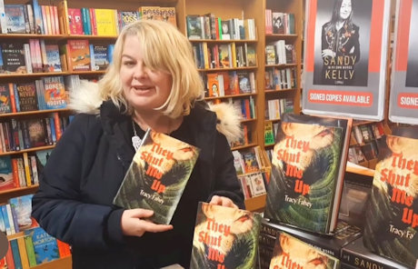 Author Tracy Fahey standing in a bookstore holding a copy of her Absinthe Books novella, They Shut Me Up, beside a display of many copies of the book