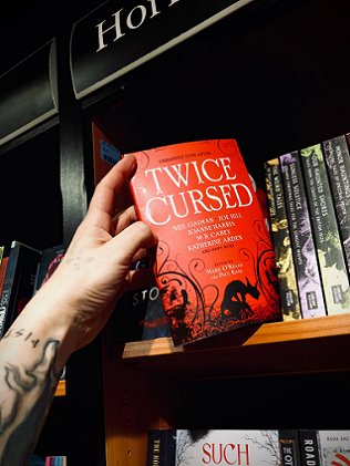Man's tattooed arm reaching out, taking a copy of Twice Cursed, edited by Marie O'Regan and Paul Kane, off a shelf in a bookstore
