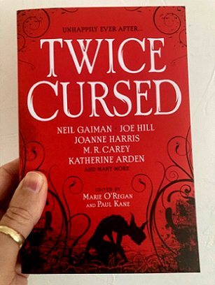 Man's hand holding a copy of Twice Cursed, edited by Marie O'Regan and Paul Kane