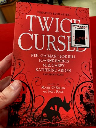 A man's hand holding a signed copy of Twice Cursed, edited by Marie O'Regan and Paul Kane