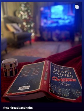 screenshot of a copy of Death Comes at Christmas, edited by Marie O'Regan and Paul Kane, face-down on a red, knitted surface, beside a white mug with toy soldiers on it. A living room decorated for Christmas is in the background, with twinkling lights on a Christmas tree, an armchair, and a TV at the back of the room