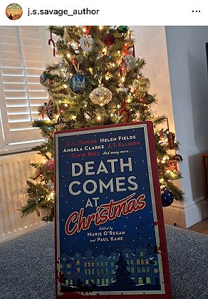 screenshot from @j.s.savage_author. Image shows a copy of Death Comes at Christmas, edited by Marie O'Regan and Paul Kane, standing against a lit and decorated Christmas tree