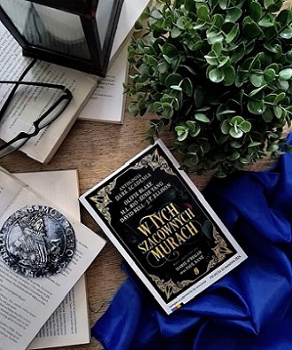 photograph showing a copy of the Polish edition of In These Hallowed Halls, edited by Marie O'Regan and Paul Kane, lying on a wooden surface beside a cobalt blue cloth near a jade plant, several open books, a pair of black-rimmed glasses and a lantern
