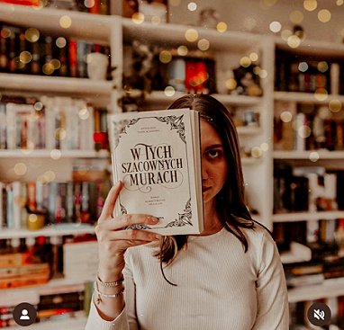 photograph of a woman with long dark hair holding up the title page of the Polish edition of In These Hallowed Halls, edited by Marie O'Regan and Paul Kane, so it obscures half her face. In the background are white bookshelves filled with books, decorated with fairy lights