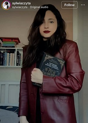 photograph of a woman with long dark hair and red lipstick, wearing a burgundy leather jacket, standing in front of bookshelves and a bed, holding a copy of the Polish edition of In These Hallowed Halls, edited by Marie O'Regan and Paul Kane.